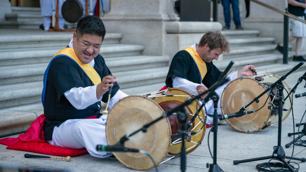 Two Samulnori percussionists performing outdoors, seated in front of their drums, on steps. Photo is from Baltimore Museum of Art.