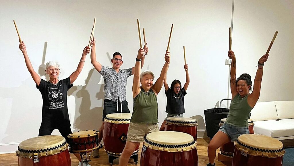 Members of BmoreTaiko (Japanese-style drumming group) standing next to their drums, with hands raised, holding drumming sticks in their hands.